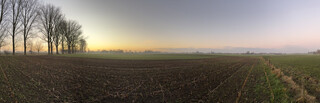 panorama-foto von einem kahlen feld, man sieht im hintergrund kahle bäume und sehr weiten, organge-blauer himmel