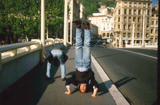 foto auf eienr br&uuml;cke in lyon, peter ist von hinten zu sehen, wie er sich b&uuml;ckt und zwischen seinen beinen in die kamera guckt, ich mache einen kopfstand und trage hosentr&auml;ger, jeans, cowboystiefel und ein jacket mit weste